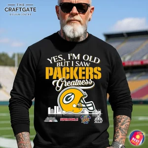 A man with a white beard and sunglasses models the black "Packers Greatness" sweatshirt in a football stadium.